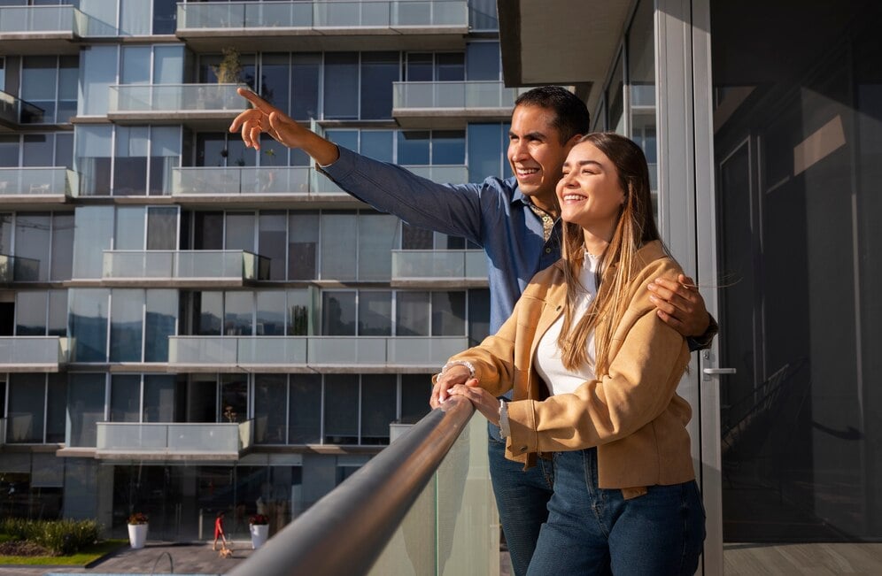 A couple standing on a balcony, pointing to another apartment