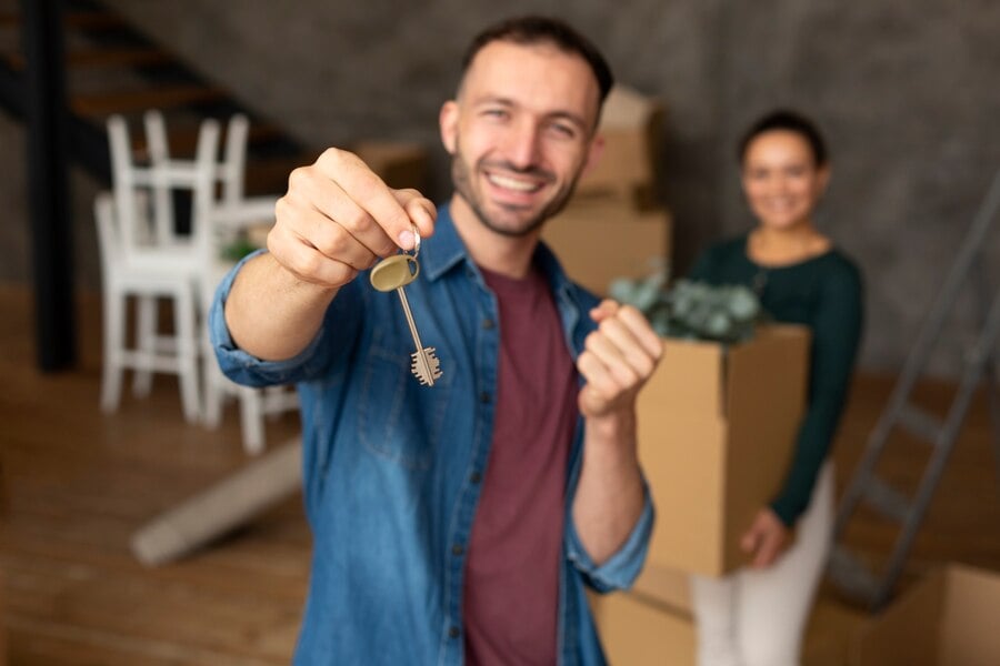 A man is holding house keys, with a woman standing in the background holding a box