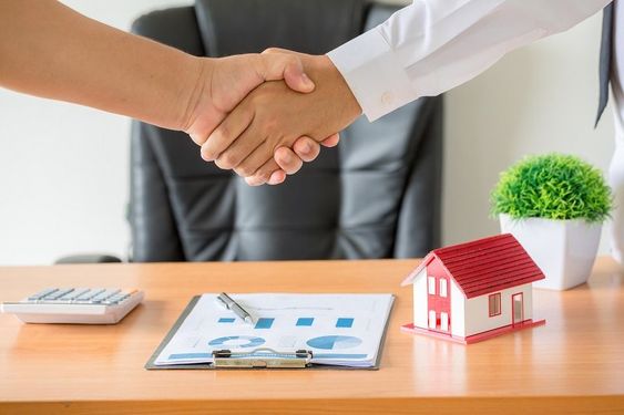 Handshake between two people with a miniature house model visible on the desk in the background