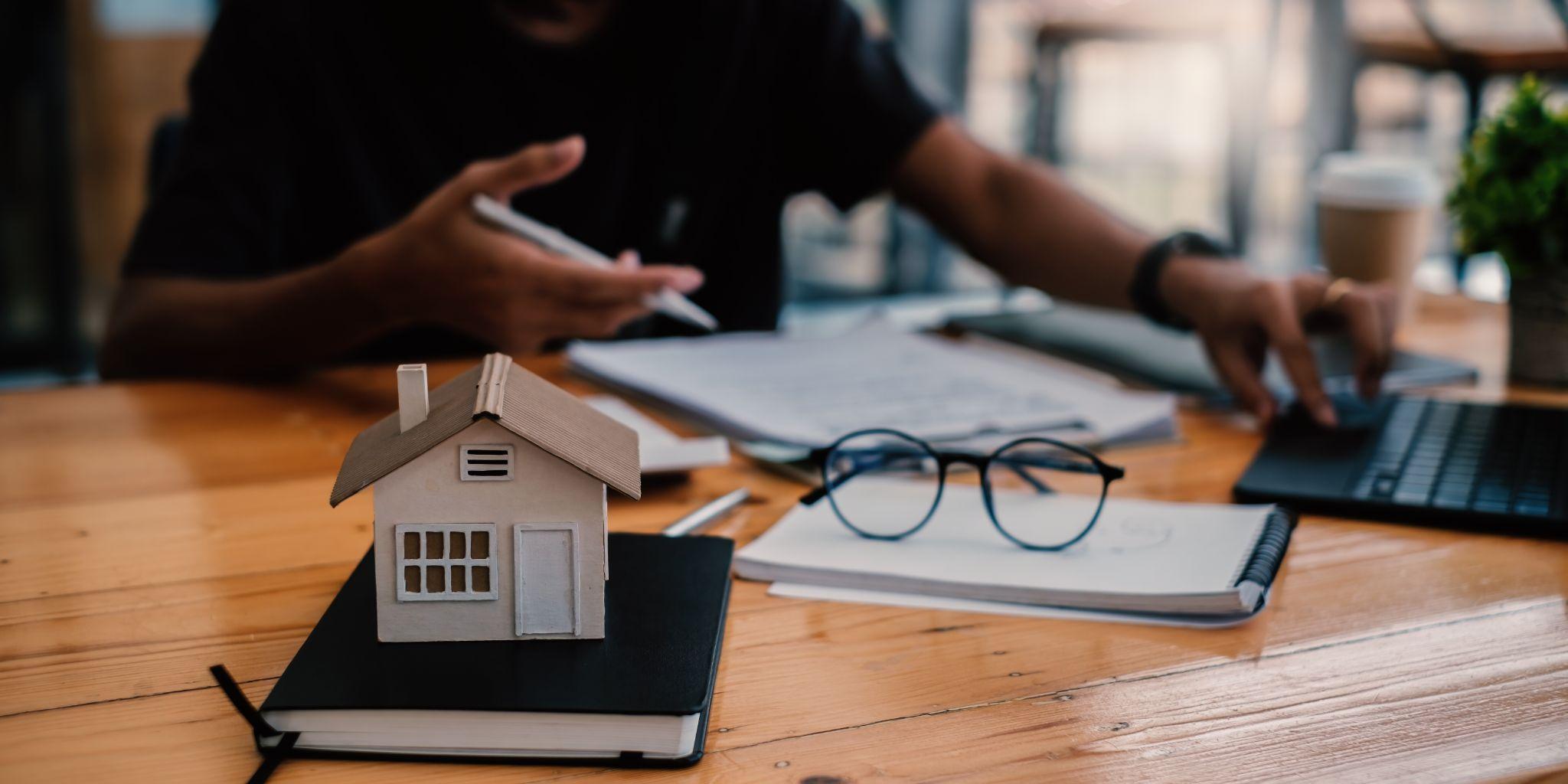 setup related to mortgages and real estate, featuring a miniature house model, eyeglasses, documents, and a person working on a laptop