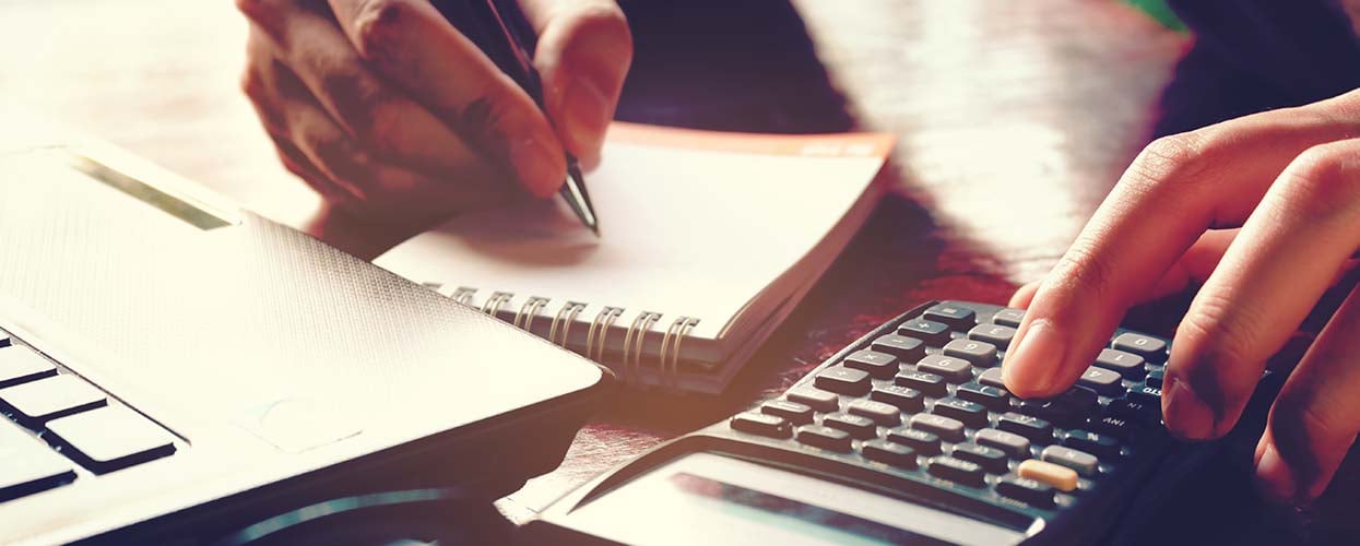 Hand writing in small notepad on table with laptop and calculator