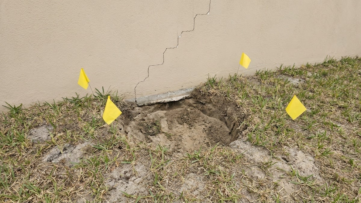 AI-generated editorial illustration of a sunken patch of soil beside a foundation slab with a stair-step crack in the stucco, not a particular property or an actual sinkhole finding.