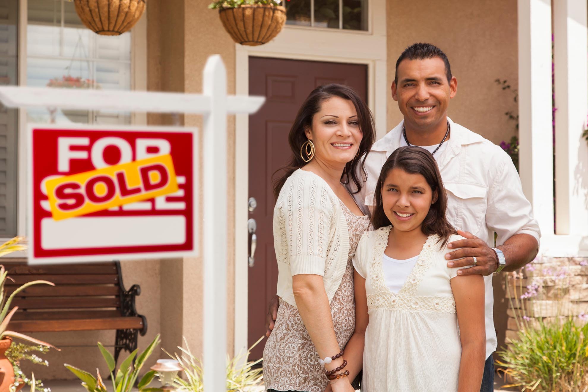 Hispanic Mother, Father and Daughter in Front of Their New Home with Sold Home For Sale Real Estate Sign.