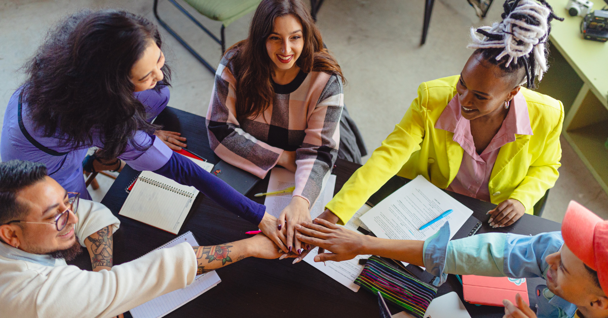 A diverse group of six professionals sitting around a table, enthusiastically participating in a team meeting and stacking hands together in unity.