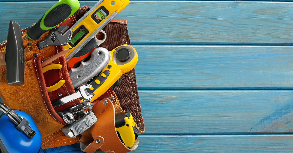 Leather tool belt filled with various hand tools, including a hammer, screwdriver, and measuring tape, on a blue wooden background.