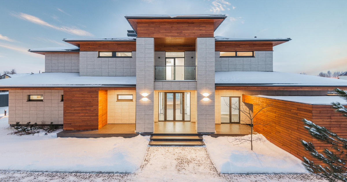 Modern two-story house with wood and stone exterior, illuminated at dusk, surrounded by snow.