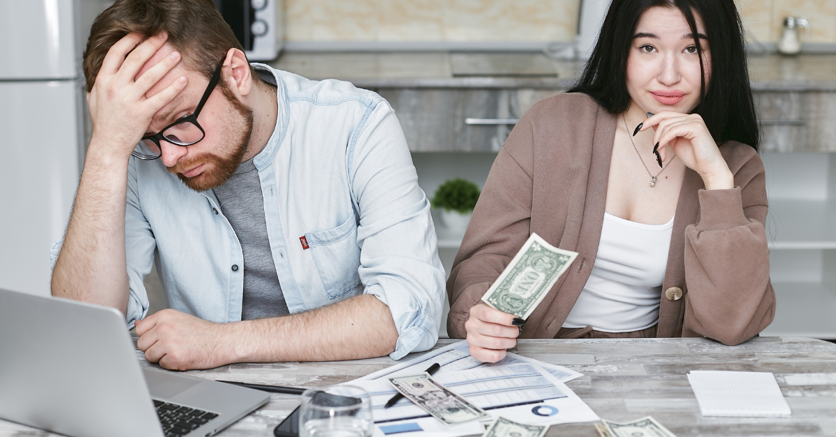 A frustrated man sitting at a table with a laptop, while a woman next to him holds a dollar bill and looks contemplative.