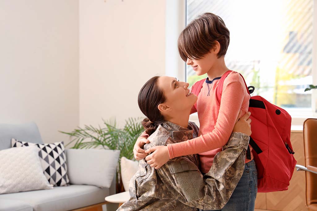 A female military personnel hugging their child, wearing a backpack, getting ready for school.