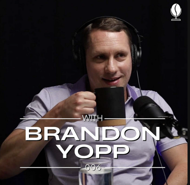 Brandon Yopp smiling and holding a black coffee mug while wearing headphones and speaking into a microphone during the recording of the Coffee & Closings podcast with Ethan Lines.