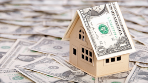 Wooden house model on a pile of US dollar bills representing home buying costs and budgeting.