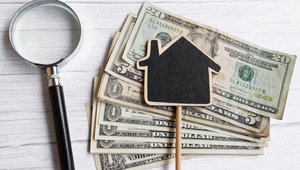 Stack of US dollar bills with a house-shaped cutout and magnifying glass representing the cost of living in St. Augustine FL.