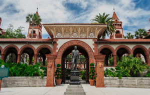 Historic Flagler College entrance in St. Augustine, Florida, featuring red-brick arches, ornate architecture, palm trees, and the statue of Henry Flagler.