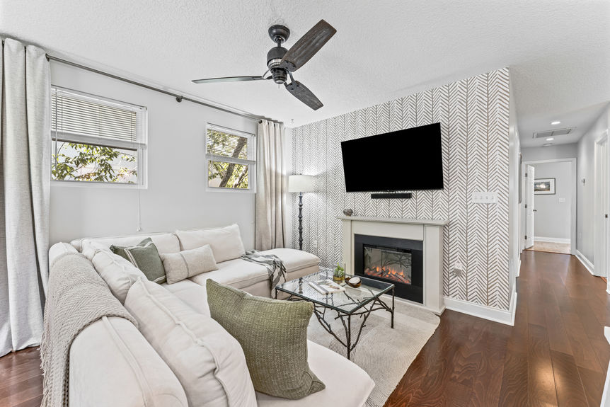 A spacious living room featuring a large white sectional sofa, a glass coffee table with branch-style legs, vaulted ceilings with a rustic fan, and a large decorative wall clock.