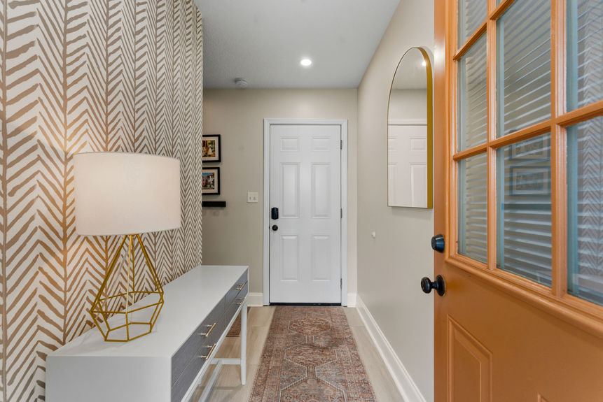 A modern home foyer featuring a patterned accent wall, a geometric gold lamp on a white console table, a decorative runner rug, and a warm wood-toned front door.