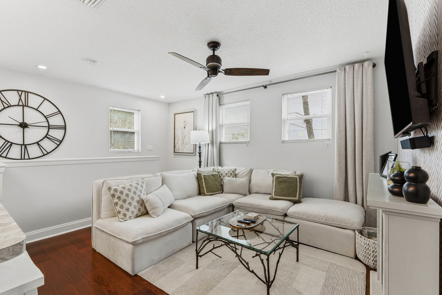 A spacious living room featuring a large white sectional sofa, a glass coffee table with branch-style legs, vaulted ceilings with a rustic fan, and a large decorative wall clock.