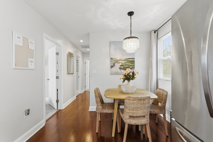 A bright dining area featuring a wooden table with four wicker chairs, a shell-style chandelier, and large windows, situated on rich hardwood floors in a St. Augustine home.