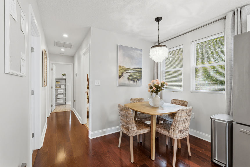 A bright dining area featuring a wooden table with four wicker chairs, a shell-style chandelier, and large windows, situated on rich hardwood floors in a St. Augustine home.