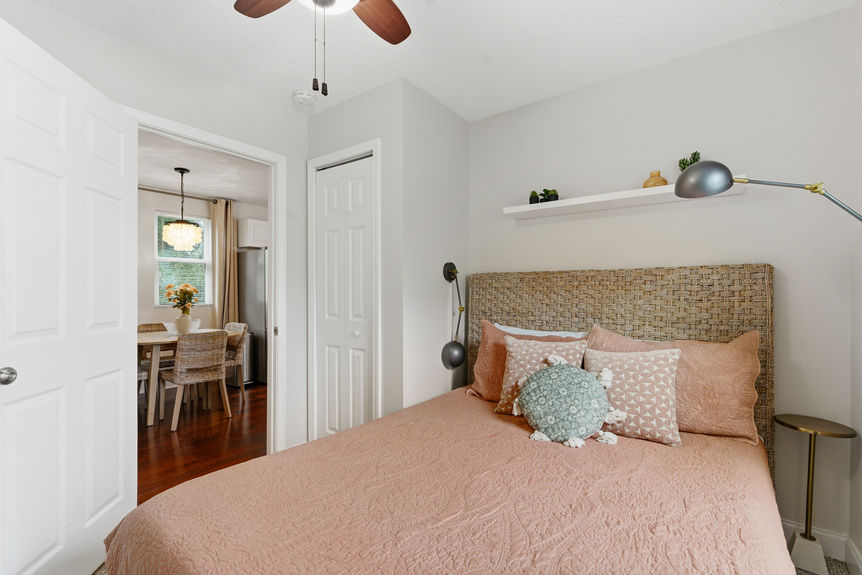A well-lit bedroom featuring a woven headboard, terracotta bedding, a wall-mounted TV, and a window with a bamboo shade overlooking St. Augustine greenery.