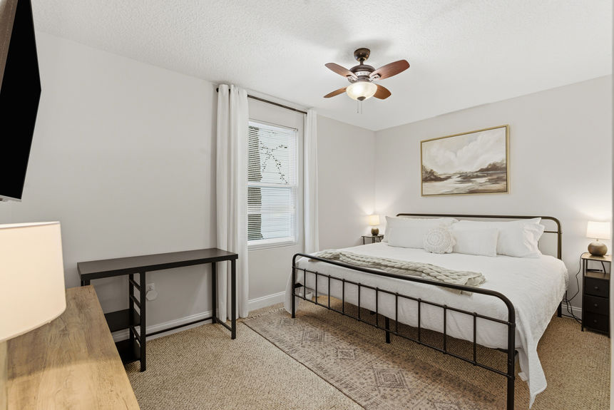 Spacious primary bedroom with a black iron bed frame, white linens, neutral wall art, and soft bedside lighting under a wood-bladed ceiling fan.