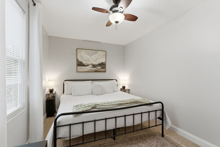 Spacious primary bedroom with a black iron bed frame, white linens, neutral wall art, and soft bedside lighting under a wood-bladed ceiling fan.