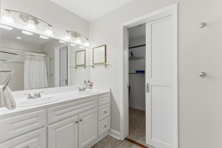 Modern bathroom with a double sink white vanity, large mirror with bell-shaped lights, and a white sliding pocket door leading into a carpeted walk-in closet.