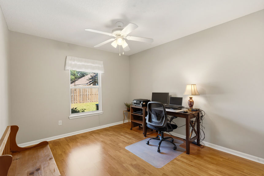 Homes for Sale in St. Johns, Florida: A bright home office or bedroom featuring hardwood-style flooring, a wooden desk with a computer setup, and a window overlooking a fenced backyard.