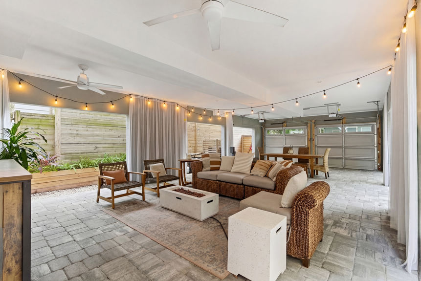 A covered outdoor lounge featuring wicker sectional seating, a modern fire pit table, privacy curtains, and string lights.