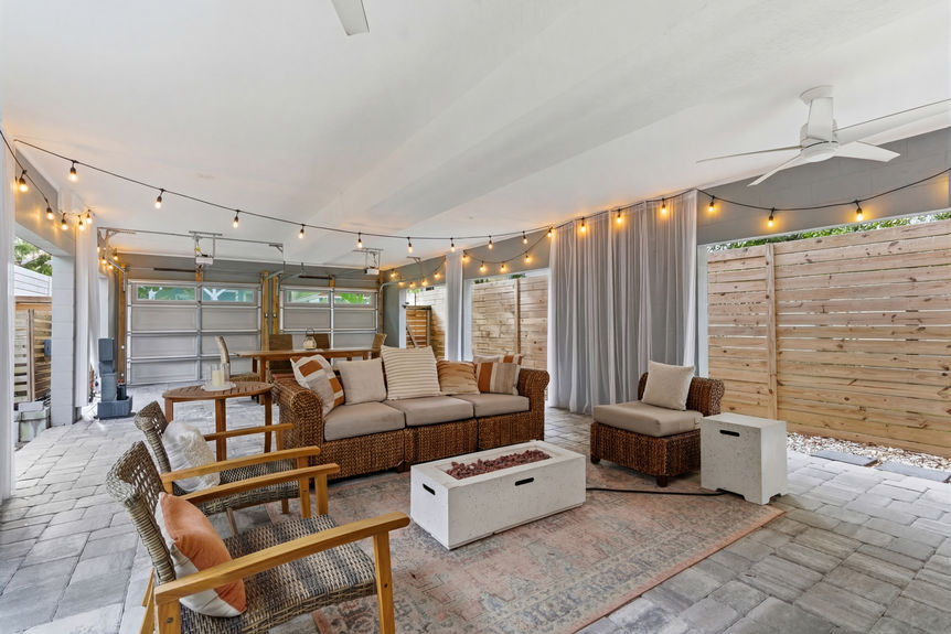 A covered outdoor lounge featuring wicker sectional seating, a modern fire pit table, privacy curtains, and string lights.