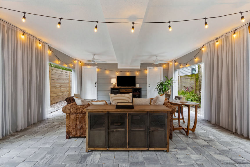 A covered outdoor lounge featuring wicker sectional seating, a modern fire pit table, privacy curtains, and string lights.