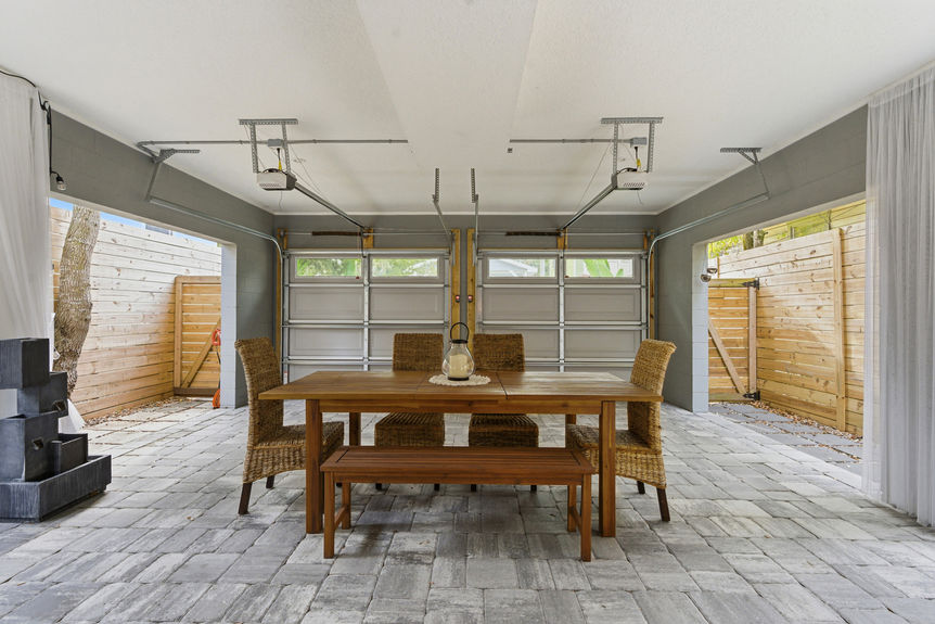 A covered outdoor lounge featuring wicker sectional seating, a modern fire pit table, privacy curtains, and string lights.