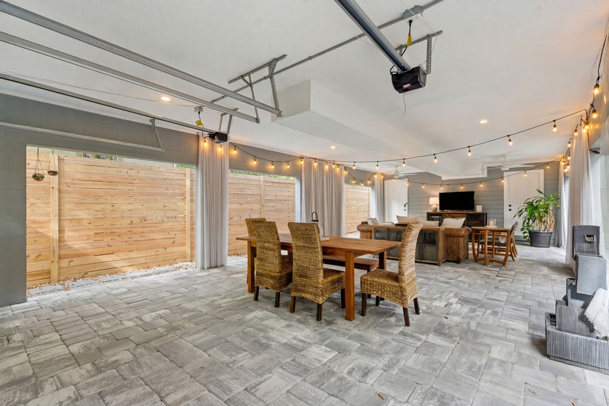 A covered outdoor lounge featuring wicker sectional seating, a modern fire pit table, privacy curtains, and string lights.