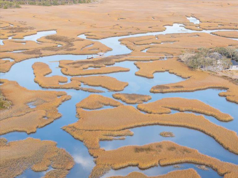 Zoomed aerial view showing a neighbor's long dock extending into the marsh, illustrating the potential lifestyle and value of the surrounding properties.