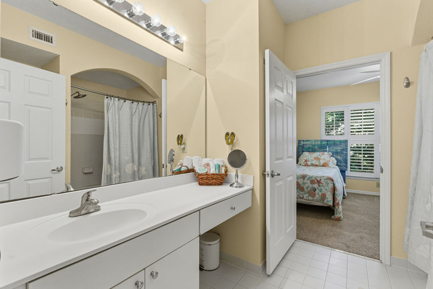 A clean bathroom featuring a long white vanity, large mirror, and a view into the bedroom.