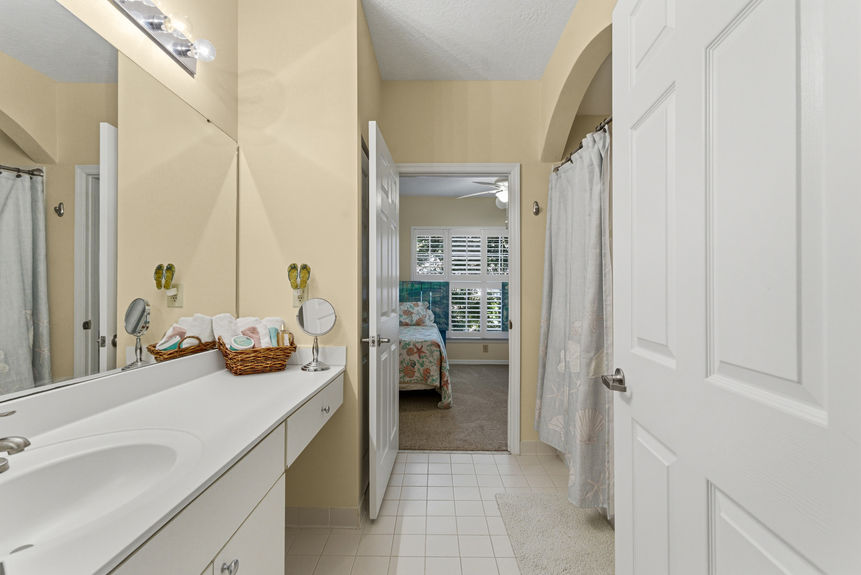 A clean bathroom featuring a long white vanity, large mirror, and a view into the bedroom.