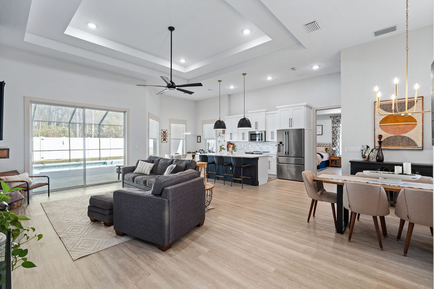 A massive great room with tray ceilings, luxury vinyl plank flooring, a chef's kitchen with a dark island, and views of the pool.