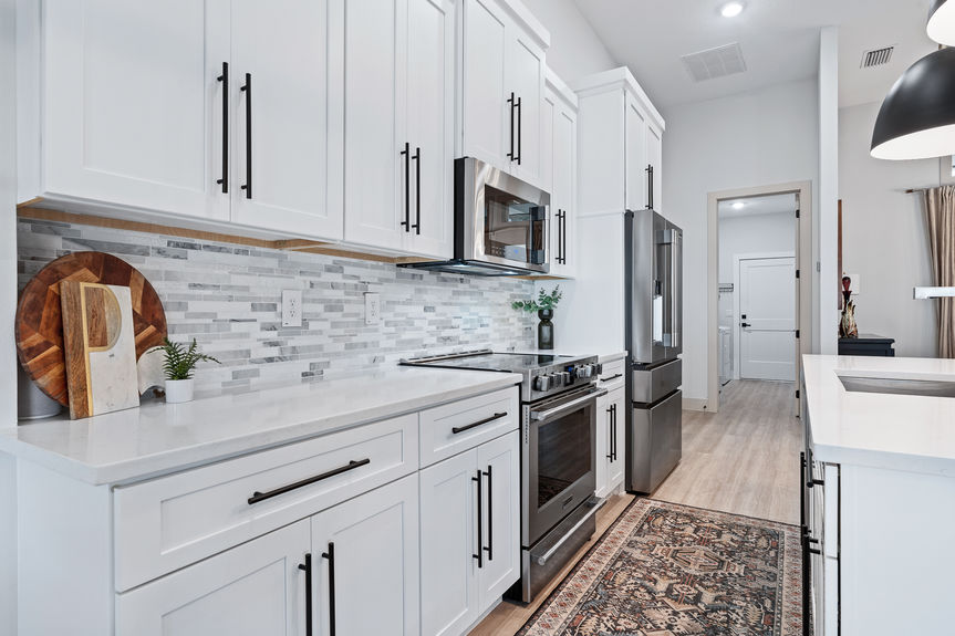 Close-up of white kitchen cabinetry, stainless steel microwave, and smooth stone countertops with a mosaic tile backsplash.