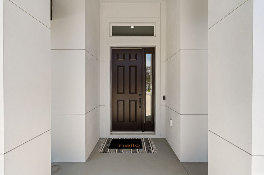 A dark, modern front door with a glass side panel and a 'hello' mat, framed by tall white pillars and a transom window.