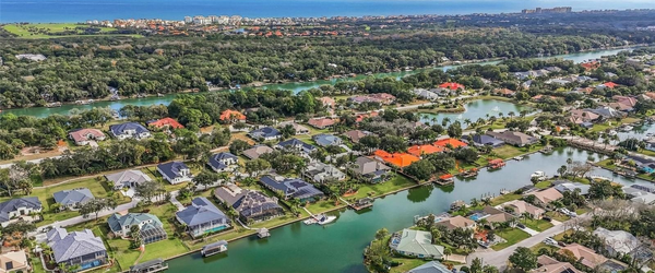 Aerial view of a waterfront neighborhood in Palm Coast, Florida, featuring luxury homes on a saltwater canal with private boat docks and a view of the Atlantic Ocean in the distance.