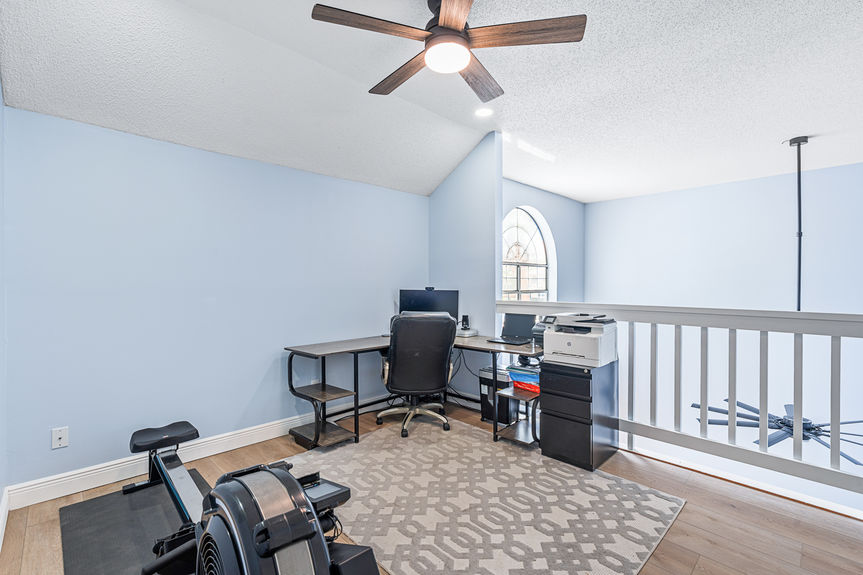 A spacious upper-level loft at 37 Captains Walk featuring a home office desk, rowing machine, and new wood-look flooring overlooking the living area.