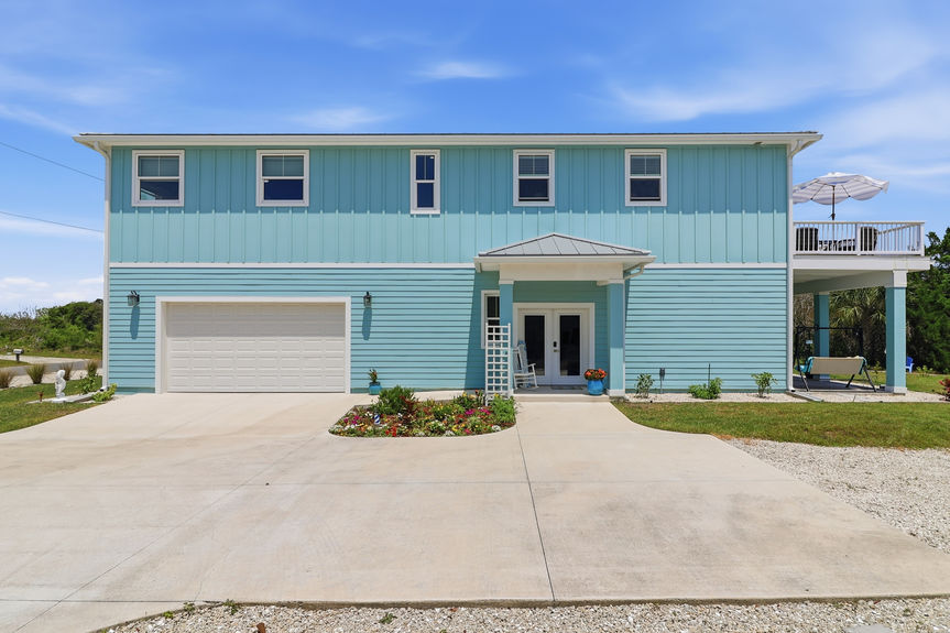 Front facade of a teal coastal house at 5900 Costanero Road in St. Augustine, FL, showing the main entryway and modern architecture under a blue sky.