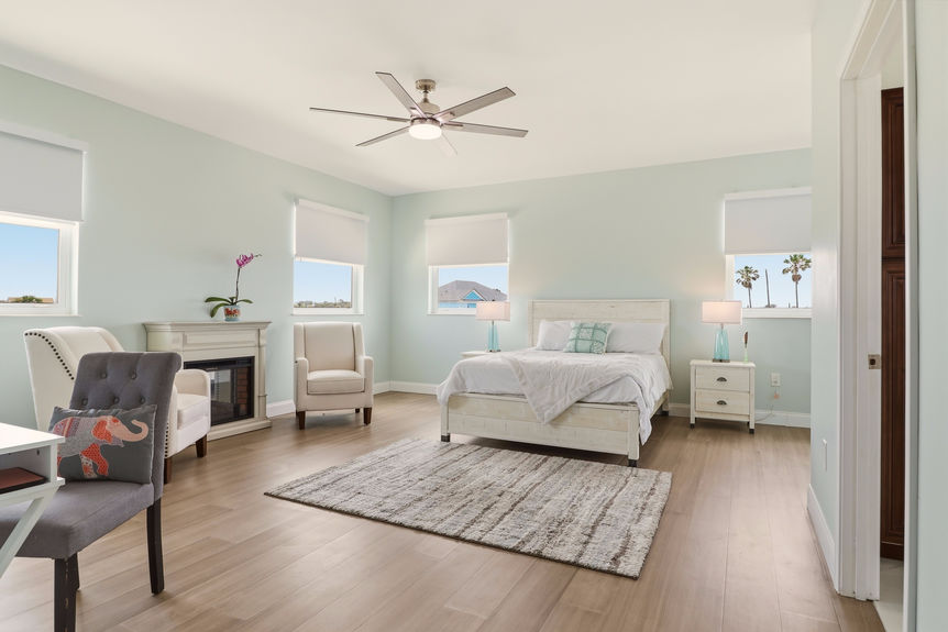 Large primary bedroom at 5900 Costanero Road with light blue walls, wood-look flooring, a sitting area with two cream armchairs, and large windows.