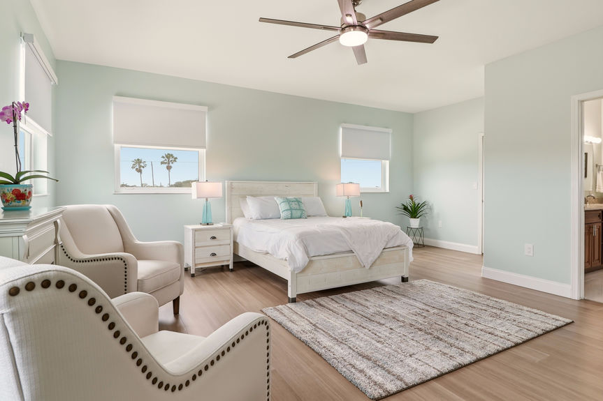 Large primary bedroom at 5900 Costanero Road with light blue walls, wood-look flooring, a sitting area with two cream armchairs, and large windows.