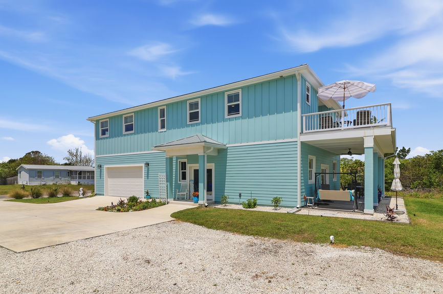 Front facade of a teal coastal house at 5900 Costanero Road in St. Augustine, FL, showing the main entryway and modern architecture under a blue sky.