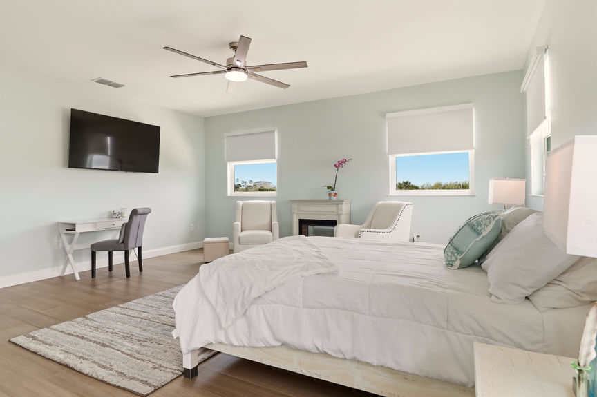 Large primary bedroom at 5900 Costanero Road with light blue walls, wood-look flooring, a sitting area with two cream armchairs, and large windows.