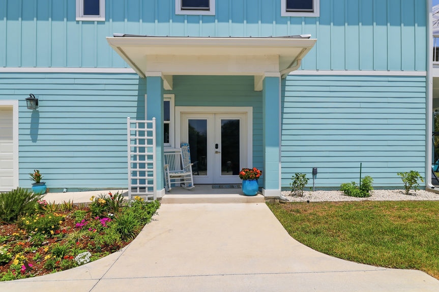 The inviting front entrance of 5900 Costanero Road featuring double glass doors, a white rocking chair, and manicured coastal landscaping.