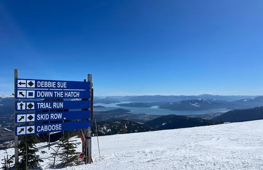 Ski trail sign at Schweitzer Mountain Resort in Sandpoint, Idaho, with a panoramic view of Lake Pend Oreille and surrounding mountains. The snow-covered slopes feature trail markers for runs like 'Debbie Sue' and 'Skid Row,' highlighting the resort's diverse terrain and stunning winter scenery.