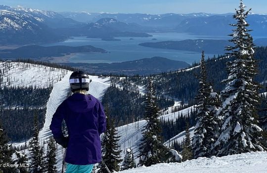 Skier at Schweitzer Mountain Resort in Sandpoint, Idaho, overlooking snow-covered slopes and a panoramic view of Lake Pend Oreille. Stunning winter landscape with evergreen trees and mountain terrain, showcasing North Idaho’s premier ski destination.