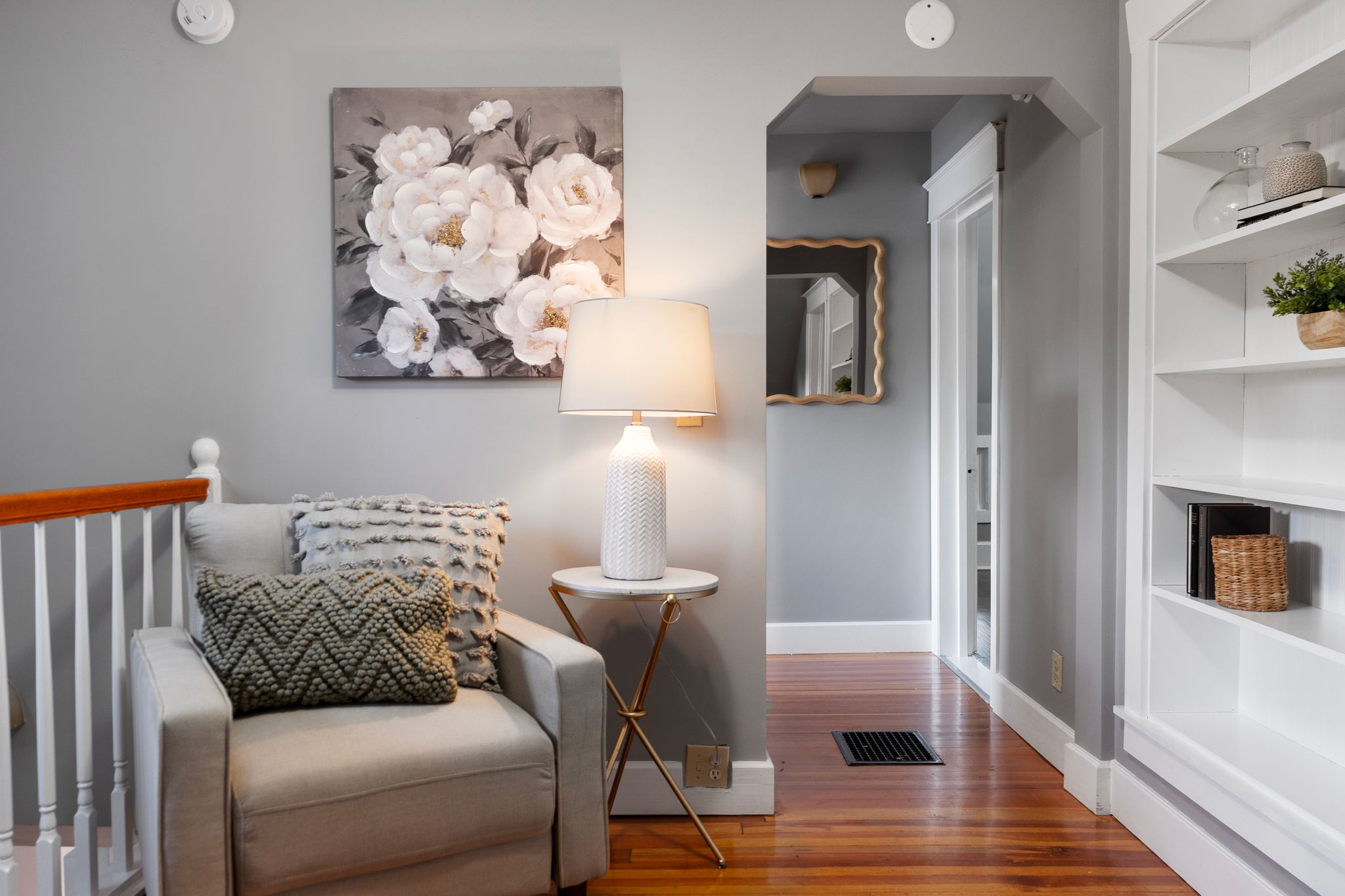 Cozy and stylish reading nook in a Sandpoint, Idaho home, featuring a neutral armchair with textured pillows, a floral wall painting, modern lamp, and built-in white shelving on hardwood floors.