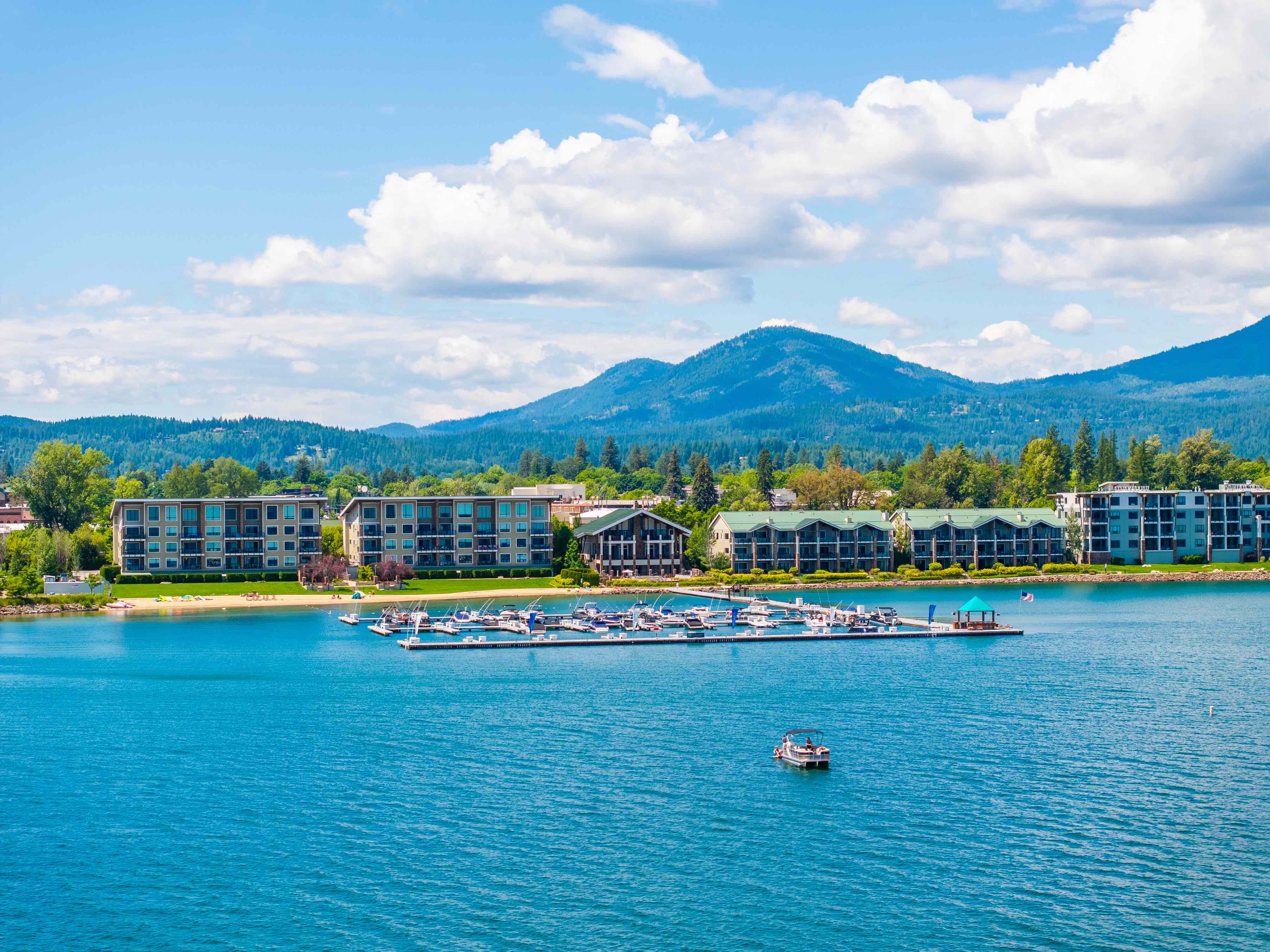 a photo of Lake Pend Oreille with The Seasons Condos, a blue sky, and mountains in the background. There is a marina with boats docked