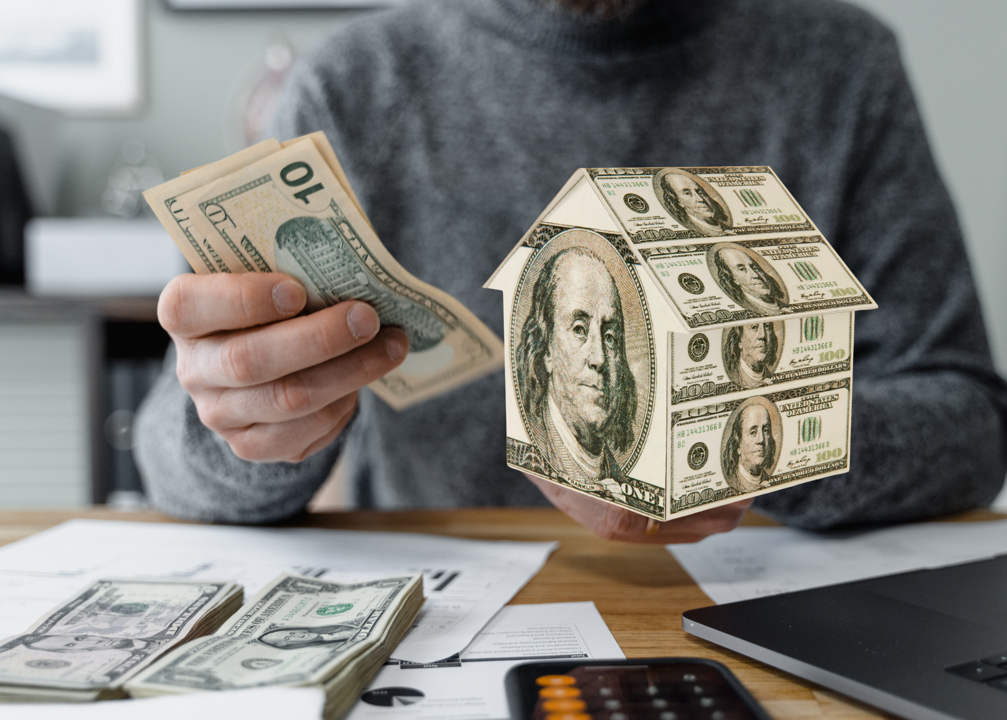 Person holding cash and a house model made of hundred-dollar bills, symbolizing real estate investment, home financing, or housing market costs